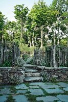 Blick auf Treppe und offenes Tor, seitlich Knüppelzaun auf Natursteinmauer vor weitläufigem Garten mit hohen Bäumen