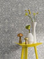 A yellow metal table with vases in front of non-woven wallpaper with a floral pattern in shades of grey