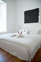 Dog on double bed with white bedspread below black painting in bedroom with wooden floor