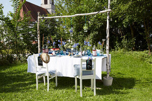 Laid table in a green setting with hanging glass vases and blue flowers