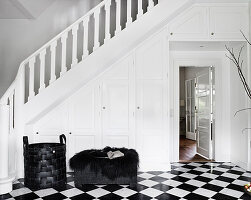 Ottoman and basket on black and white tiled floor in the hallway, white built-in cupboards under the stairs