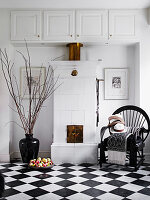 Black lacquered chair next to tiled stove, fruit bowl and floor vase on a black and white tiled floor