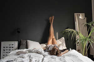 Junge Frau liegt auf Doppelbett, Beine in die Höhe gestreckt, vor grauer Wand