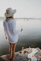 Frau im Strandkleid mit Sektglas am Felsufer eines Sees