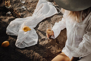 Woman wearing beach dress and sun hat holding glass of sparkling wine on rocky lake shore