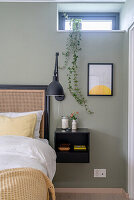 Bedside cabinet, wall lamp, hanging plant above on windowsill in bedroom