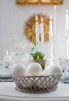 Metallkorb mit weißen Eiern, im Hintergrund brennende Kerzen auf Ostertisch