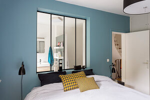 Double bed in front of an interior window with view of the bathroom in a bedroom with a blue wall