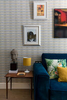 Sofa mit blauem Samtbezug und Kissen, Beistelltisch mit Buddha und gemusterte Tapete mit Foto und Poster