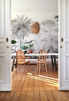 Table with various chairs and houseplants in dining room with mural wallpaper and wooden floorboards