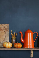 Orange enamel coffee pot on a shelf against a dark grey wall with pumpkins candles and a wooden board