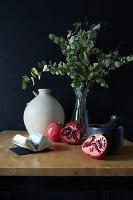 Tisch mit Mörser, Granatäpfeln, Vase und Eukalyptuszweigen vor schwarzer Wand
