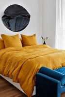 An apartment bedroom with abstract acrylic art, turmeric-coloured crushed linen bedding and a dark blue velvet bench