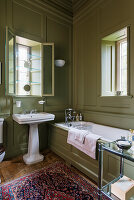 Olivgrün getäfeltes Badezimmer mit Glasregalen in der Fensternische und antikem Teppich