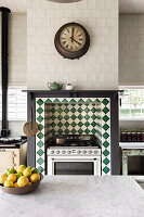 View over kitchen island with marble worktop to gas cooker in wall niche with green-white tiles, above kitchen clock