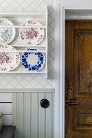 Plate rack on wallpapered wall, wood panelling underneath, next to rustic wooden door