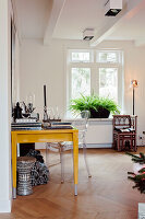 Heller Arbeitsbereich mit gelbem Schreibtisch, transparentem Stuhl und Grünpflanze am Fenster