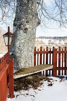 Holzbank um Baumstamm mit rotem Holzzaun und Laterne im verschneiten Garten