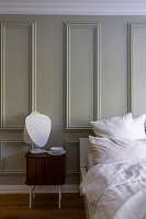 Wooden bedside table with table lamp, wall panelling in pastel green and bed with pillows and blanket