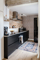 Black fitted kitchen with brick wall, patterned wallpaper and oriental carpet