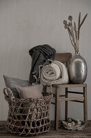 Basket with cushions, woolen blankets and dried flowers in metal vase on wooden chair