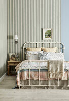 Metal bed with patterned cushions and blankets and striped wallpaper in the bedroom