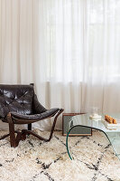 Leather chair and glass table on deep-pile carpet in front of light-coloured curtains