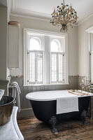 Freestanding black bathtub on wooden floorboards with chandelier and louvre windows