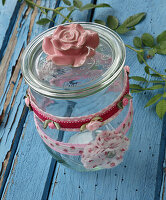 Self-sewn ribbons made of rose fabric, mini roses and fabric rosettes as decoration for storage jar and glass lid with rose on blue wooden table